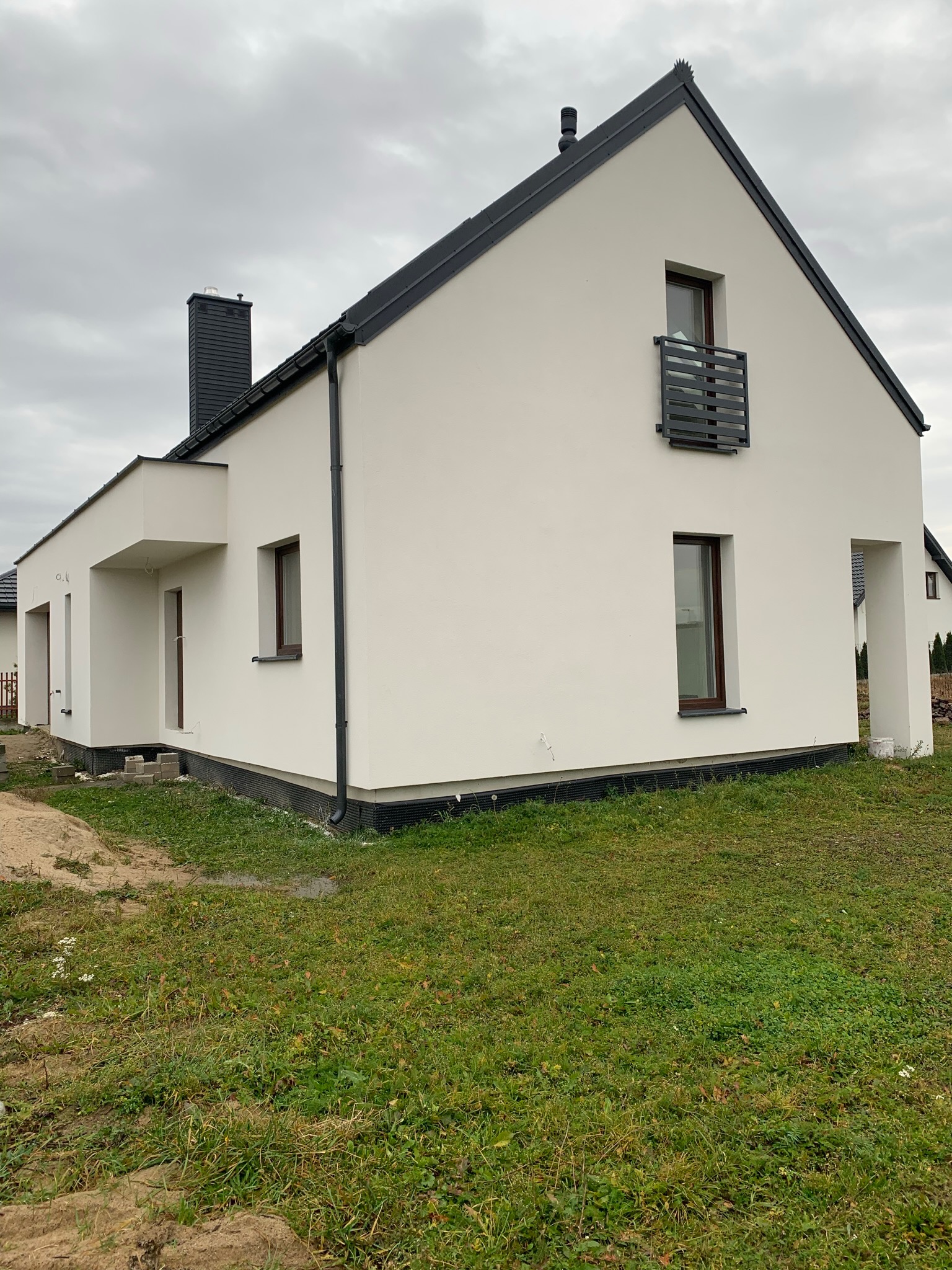 Nowoczesny, ocieplony dom jednorodzinny z białymi ścianami, ciemną dachówką i czarnymi rynnami, widoczny z boku na tle pochmurnego nieba.