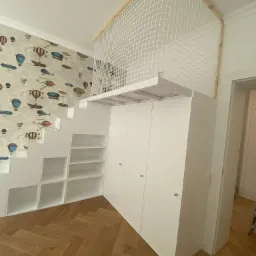 Biała antresola z siatką zabezpieczającą, wbudowana nad szafą i schodami z półkami, na tle ściany z tapetą w balony i samoloty.