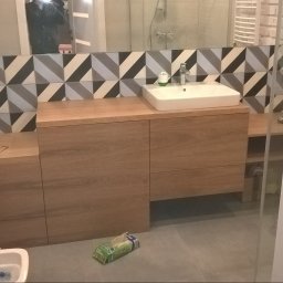 ARCHANS ARKADIUSZ KANIA - Nowoczesna łazienka z drewnianą zabudową umywalki, geometrycznymi płytkami na ścianie i szklaną kabiną prysznicową w tle.