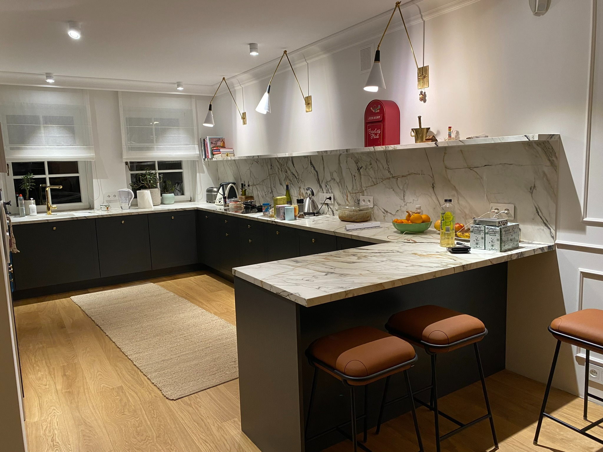 Nowoczesna kuchnia z ciemnymi szafkami, blatem imitującym marmur, oświetleniem punktowym i dekoracyjnymi lampami ściennymi; widoczne brązowe hokery i dywan na drewnianej podłodze.