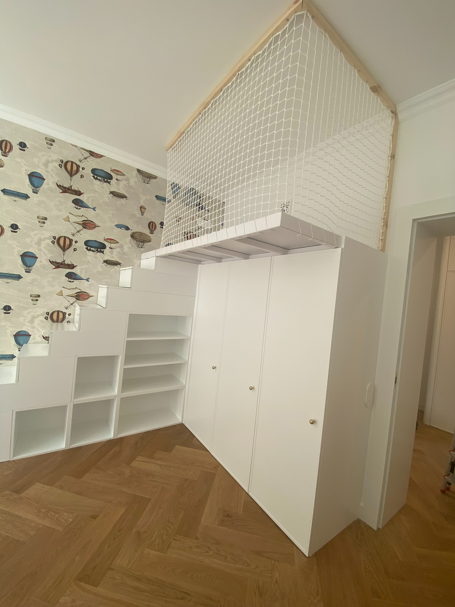 Biała antresola z siatką zabezpieczającą, wbudowana nad szafą i schodami z półkami, na tle ściany z tapetą w balony i samoloty.