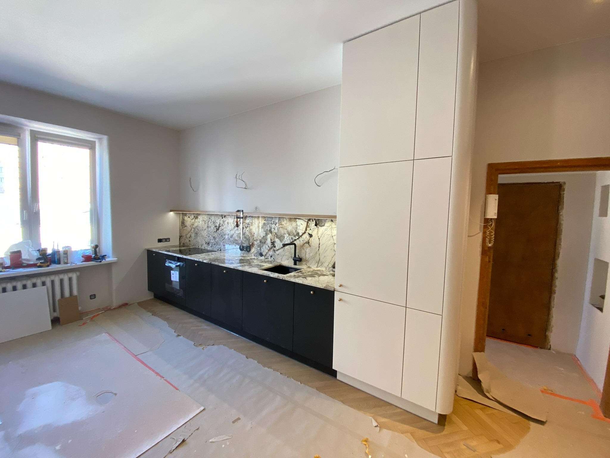 Montaż kuchni: Czarny dół, biała góra, marmurowy backsplash, widoczne niedokończone podłogi i narzędzia, sugerujące etap prac wykończeniowych.