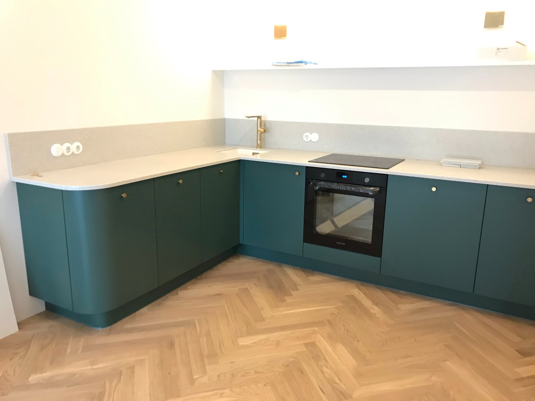 Nowoczesna kuchnia z zielonymi szafkami, jasnym blatem i drewnianą podłogą w jodełkę; widoczny piekarnik i złota bateria.