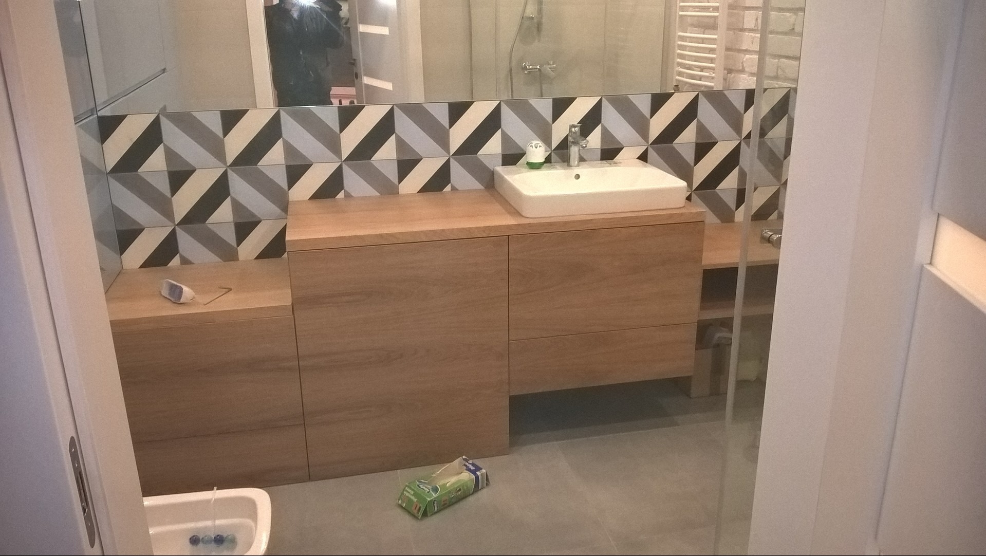 Nowoczesna łazienka z drewnianą zabudową umywalki, geometrycznymi płytkami na ścianie i szklaną kabiną prysznicową w tle.