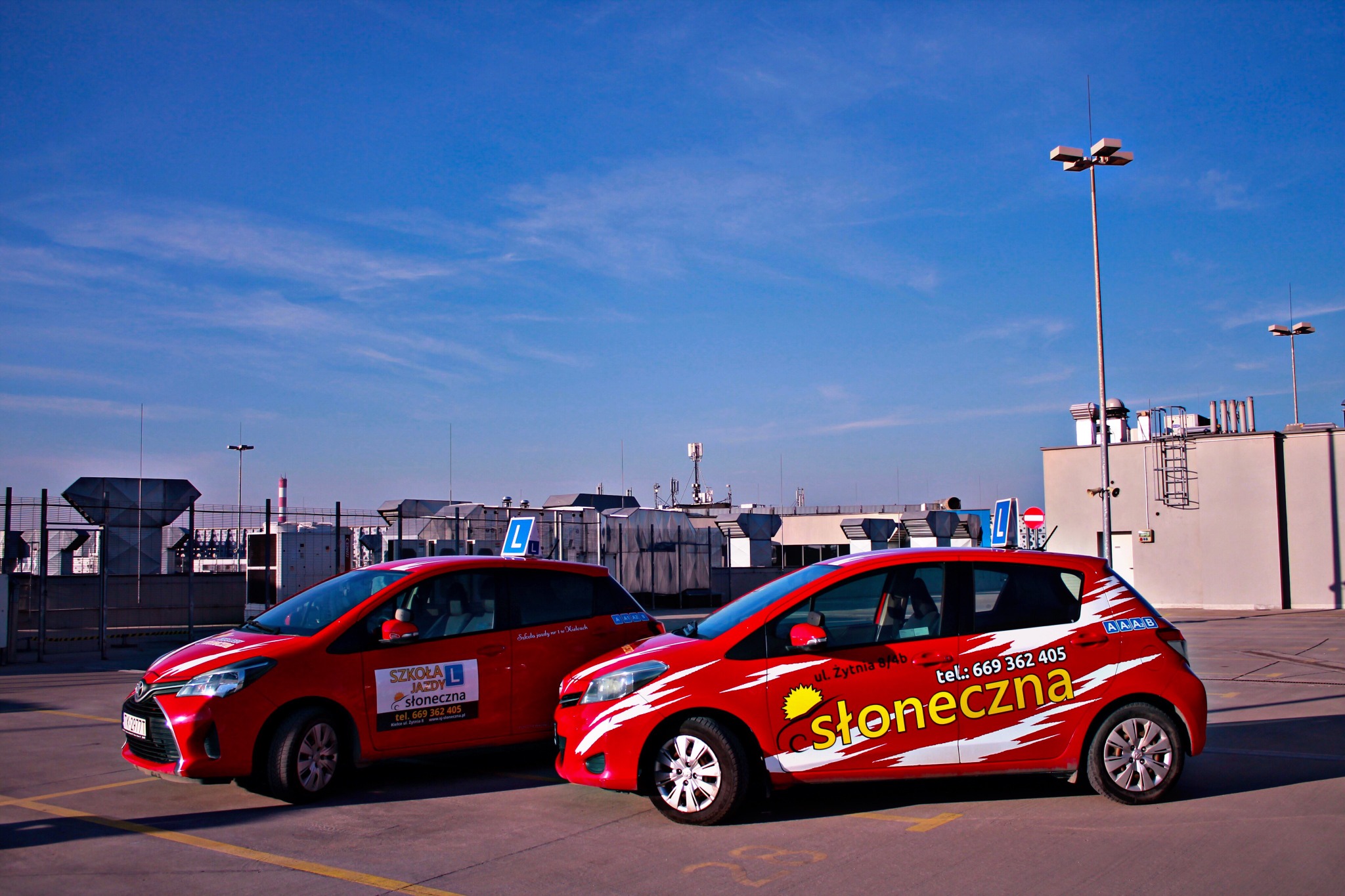 Dwa czerwone samochody z oznaczeniami nauki jazdy, zaparkowane na parkingu zewnętrznym z widocznymi budynkami przemysłowymi w tle i błękitnym niebem.
