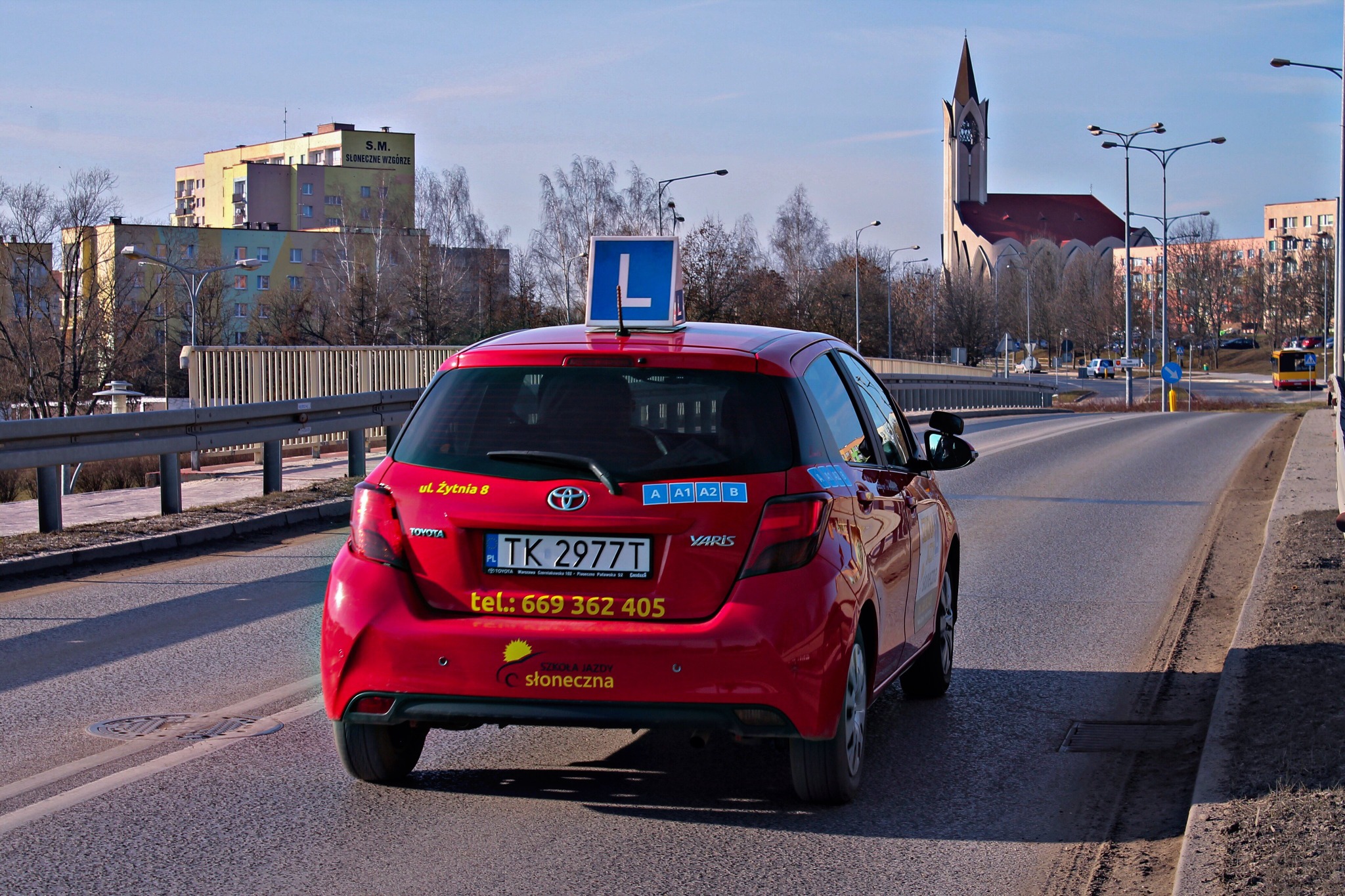 Czerwony samochód Toyota Yaris z niebieską literą 'L' na dachu, naklejki z kategoriami prawa jazdy A, A1, A2, B, numerem telefonu i adresem na tylnej klapie, jadący po asfalcie w miejskim...