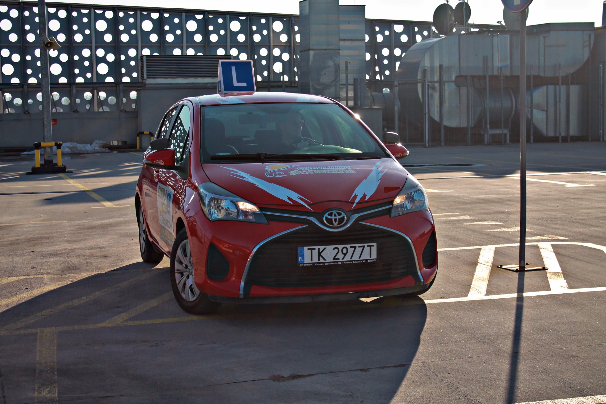 Czerwony samochód z literą 'L' na dachu, zaparkowany na placu manewrowym, widoczne oznaczenia poziome i nowoczesna architektura w tle.