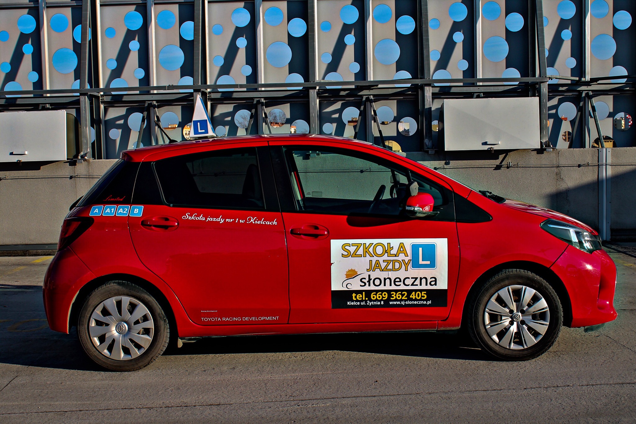 Czerwony samochód z oznaczeniami kursanta nauki jazdy, zaparkowany na tle metalowej konstrukcji z okrągłymi otworami, widok z boku.