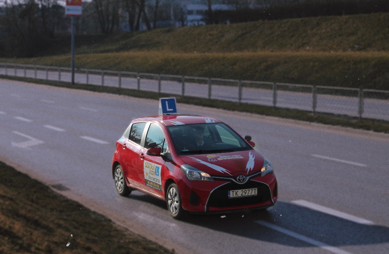Czerwony samochód Toyota Yaris z oznaczeniem 'L' na dachu, należący do szkoły jazdy, jadący po asfaltowej drodze w słoneczny dzień.