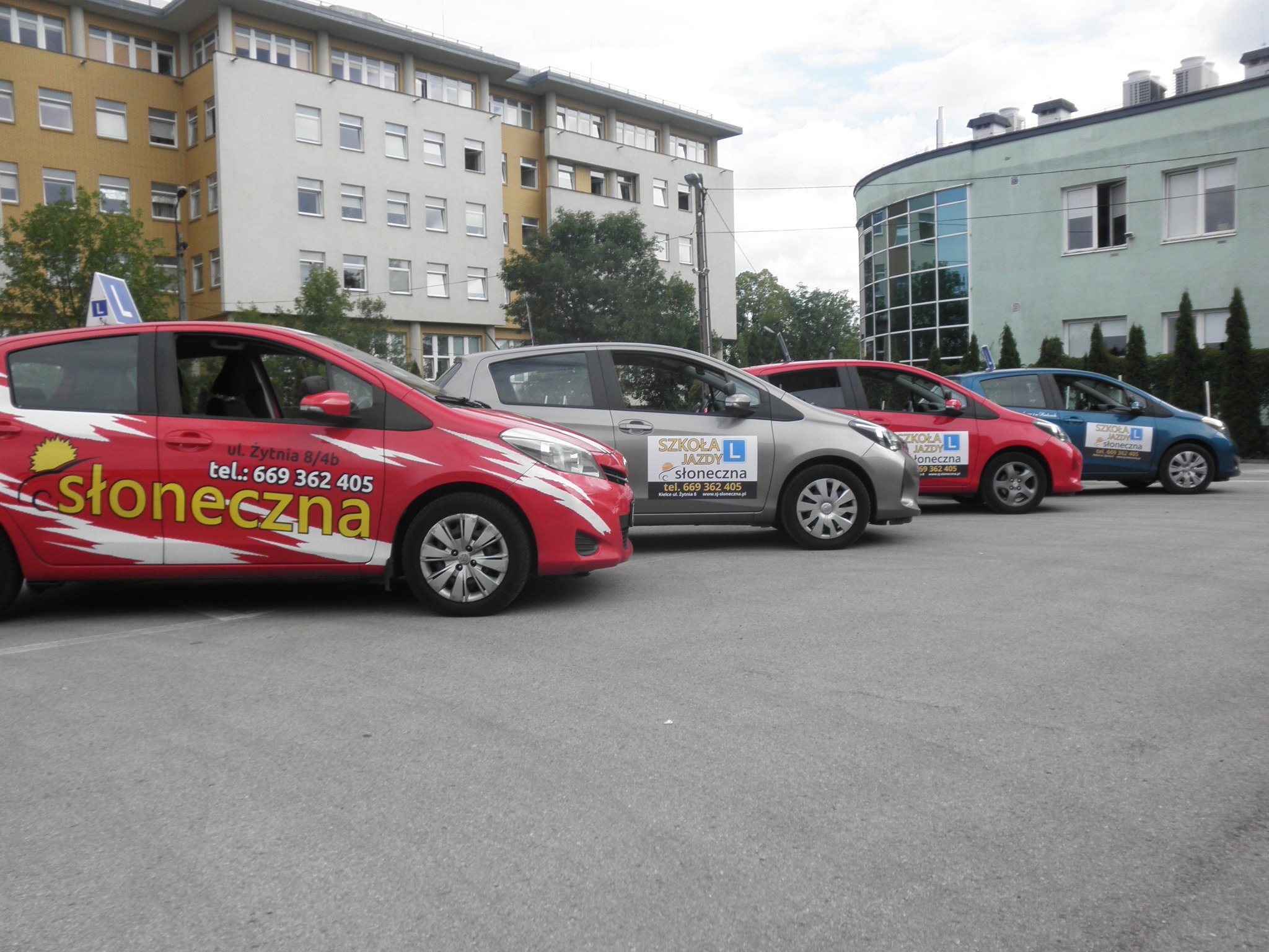 Flota samochodów z oznaczeniami nauki jazdy zaparkowana na placu, widoczne budynki w tle.
