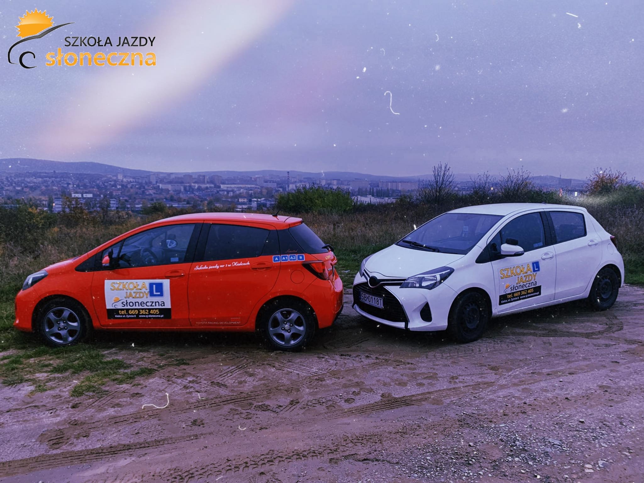 Dwa samochody Toyota Yaris, jeden pomarańczowy, drugi biały, z logo firmy z Kielc oferującej kursy nauki jazdy, zaparkowane na nierównym terenie z widokiem na panoramę miasta w tle.