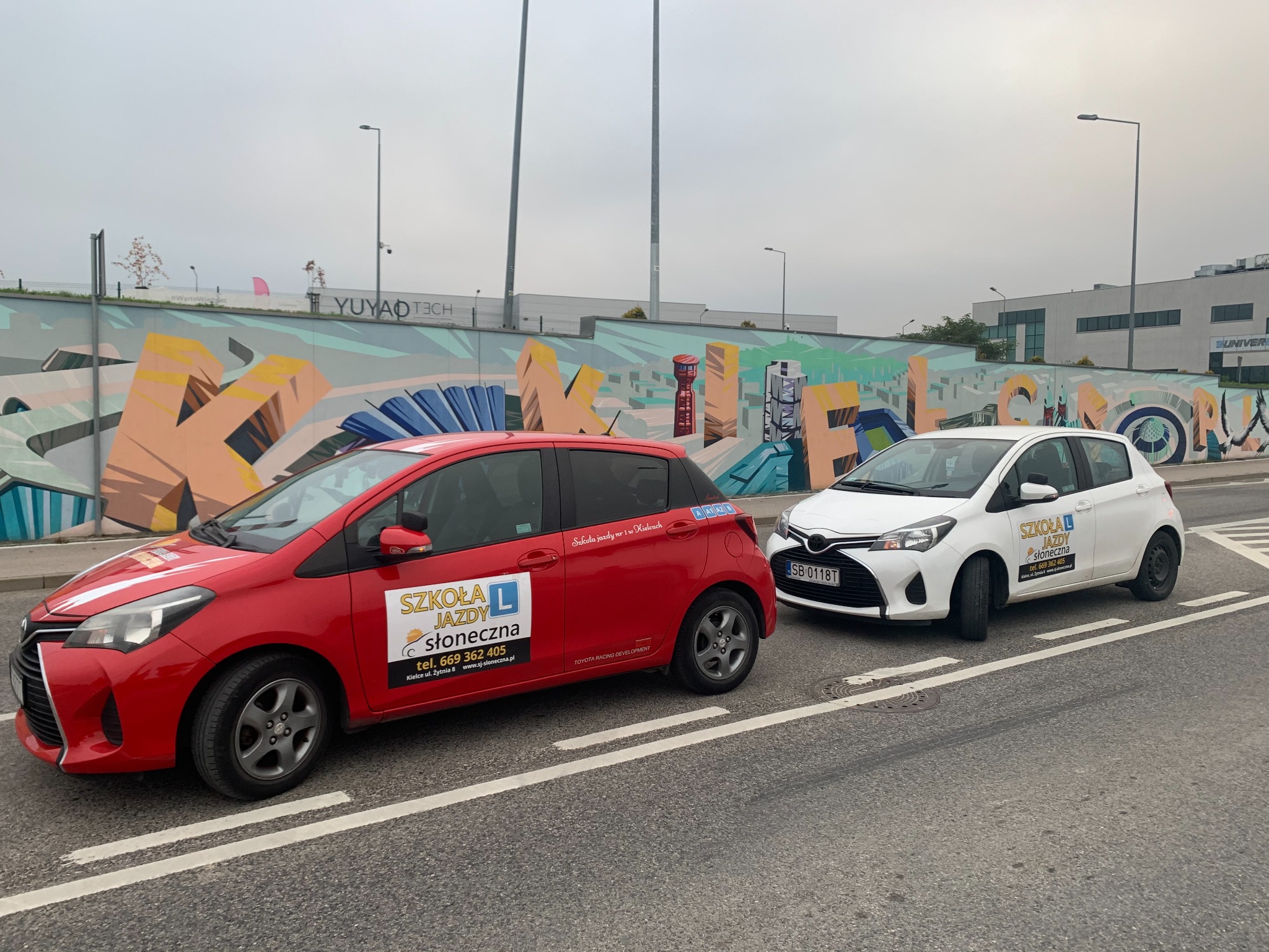 Dwa samochody z oznaczeniami firmy nauki jazdy, czerwony i biały, zaparkowane na ulicy na tle muru z kolorowym graffiti.