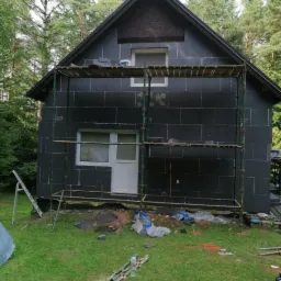 Dom jednorodzinny w trakcie ocieplania elewacji, pokryty czarnymi płytami izolacyjnymi, otoczony rusztowaniem, z widocznymi narzędziami i materiałami budowlanymi na trawniku.