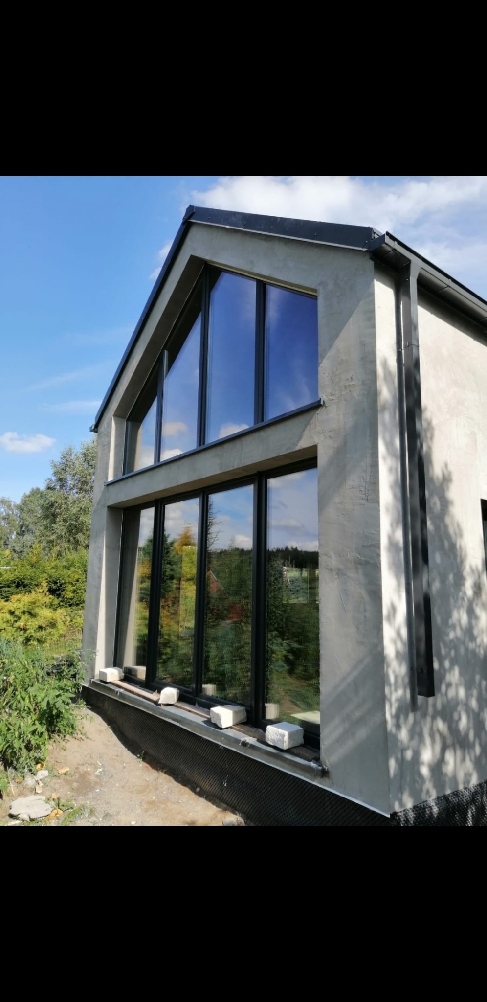 Surowa elewacja domu szkieletowego z dużymi oknami, widoczne białe bloczki pod oknami i fragment ogrodu w tle.