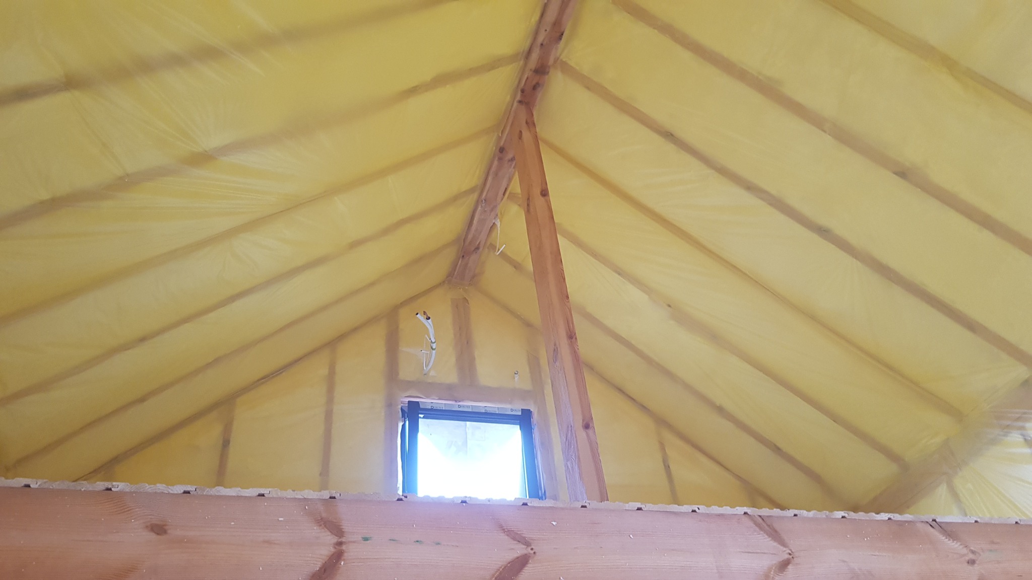 Poddasze z oknem dachowym, ocieplone pianką PUR, widoczne krokwie i elementy drewniane konstrukcji dachu, folia ochronna na piance.