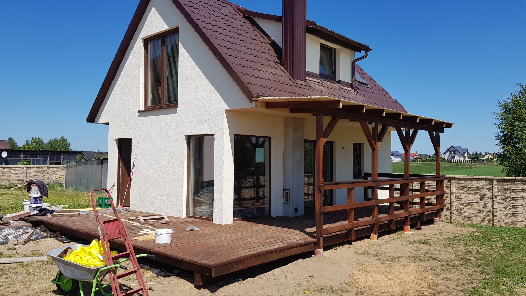 Nowoczesny dom jednorodzinny z brązowym dachem i drewnianym tarasem w trakcie budowy, widoczne narzędzia i materiały budowlane, jasne elewacje w słoneczny dzień.