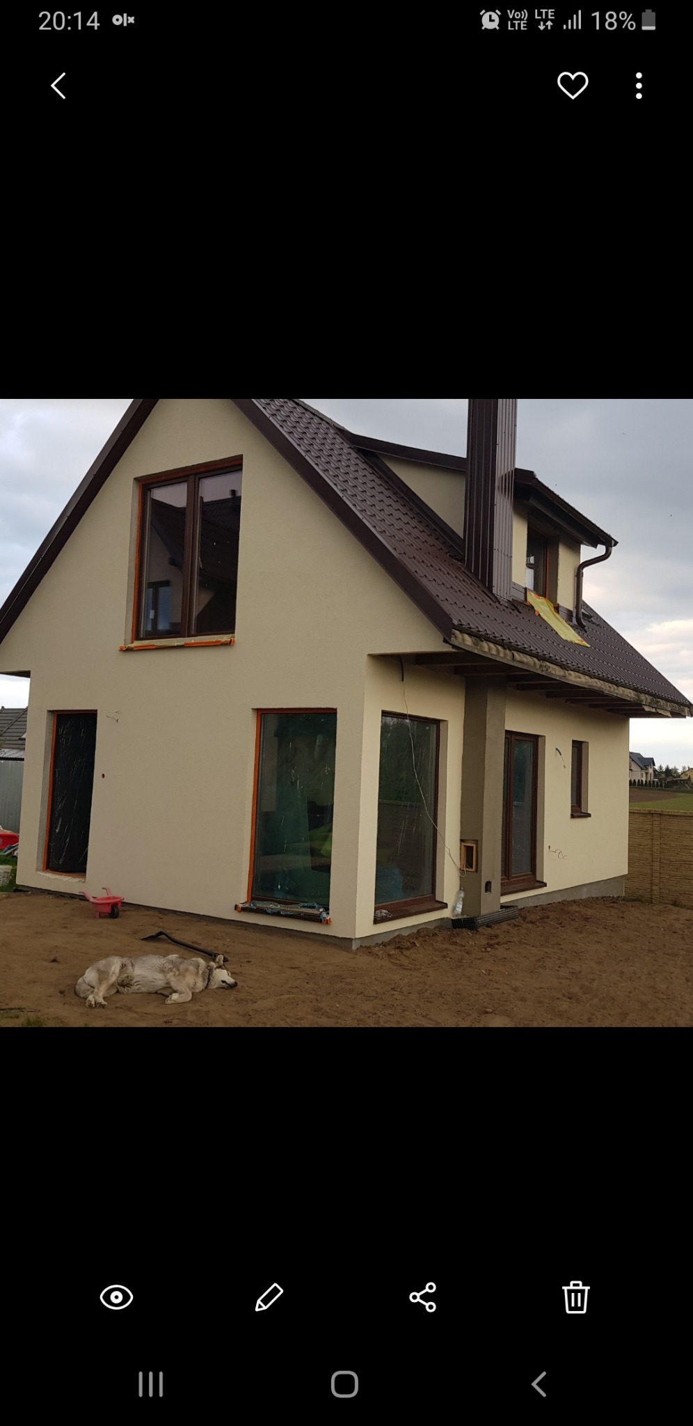 Jasny dom jednorodzinny z brązowym dachem i ramami okiennymi po tynkowaniu, z psem leżącym na ziemi przed budynkiem.