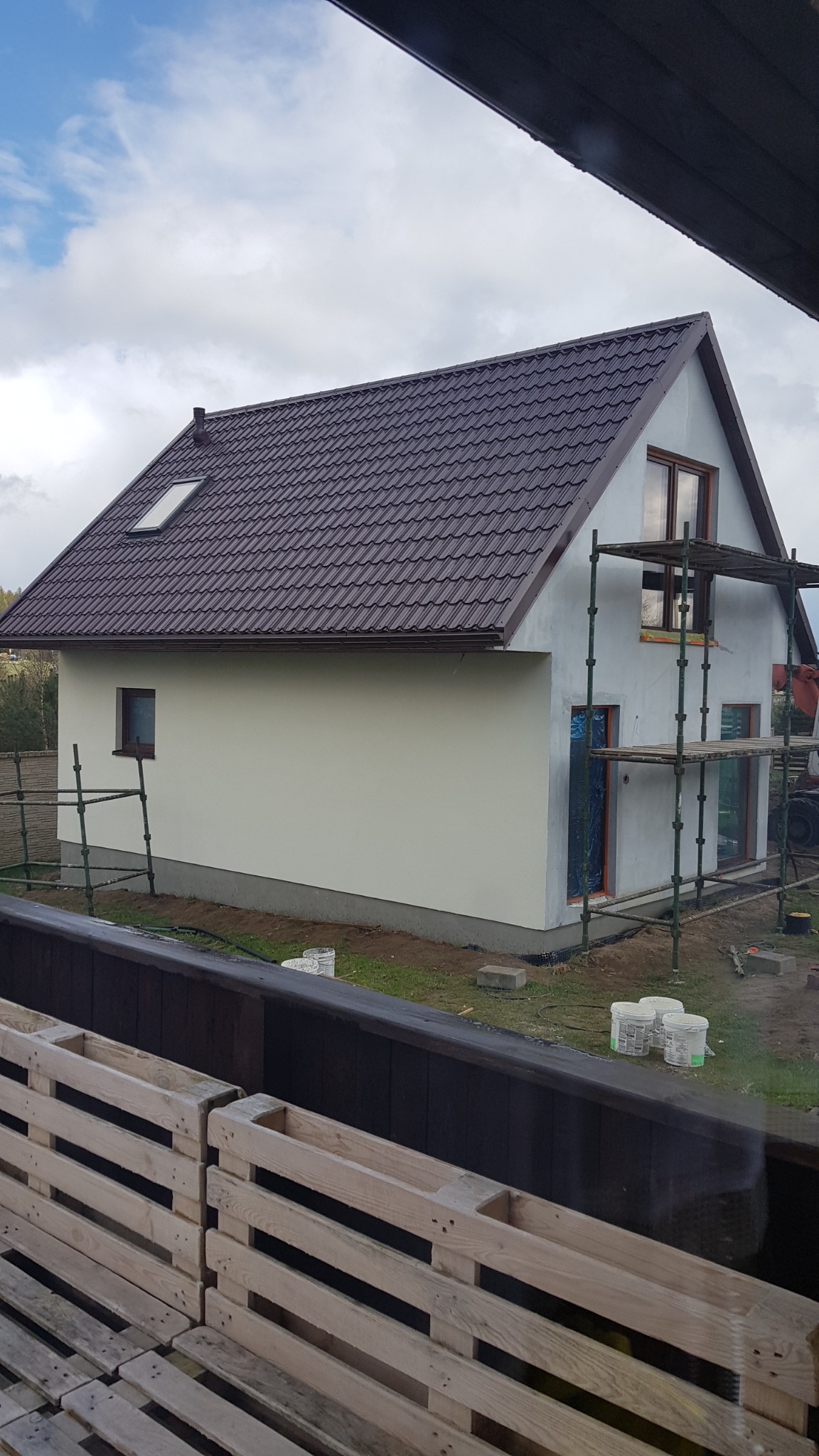 Nowo wybudowany dom jednorodzinny z brązowym dachem i jasną elewacją w trakcie wykończenia, widoczne rusztowania i farby w wiaderkach na trawniku, ujęcie zza drewnianej konstrukcji.