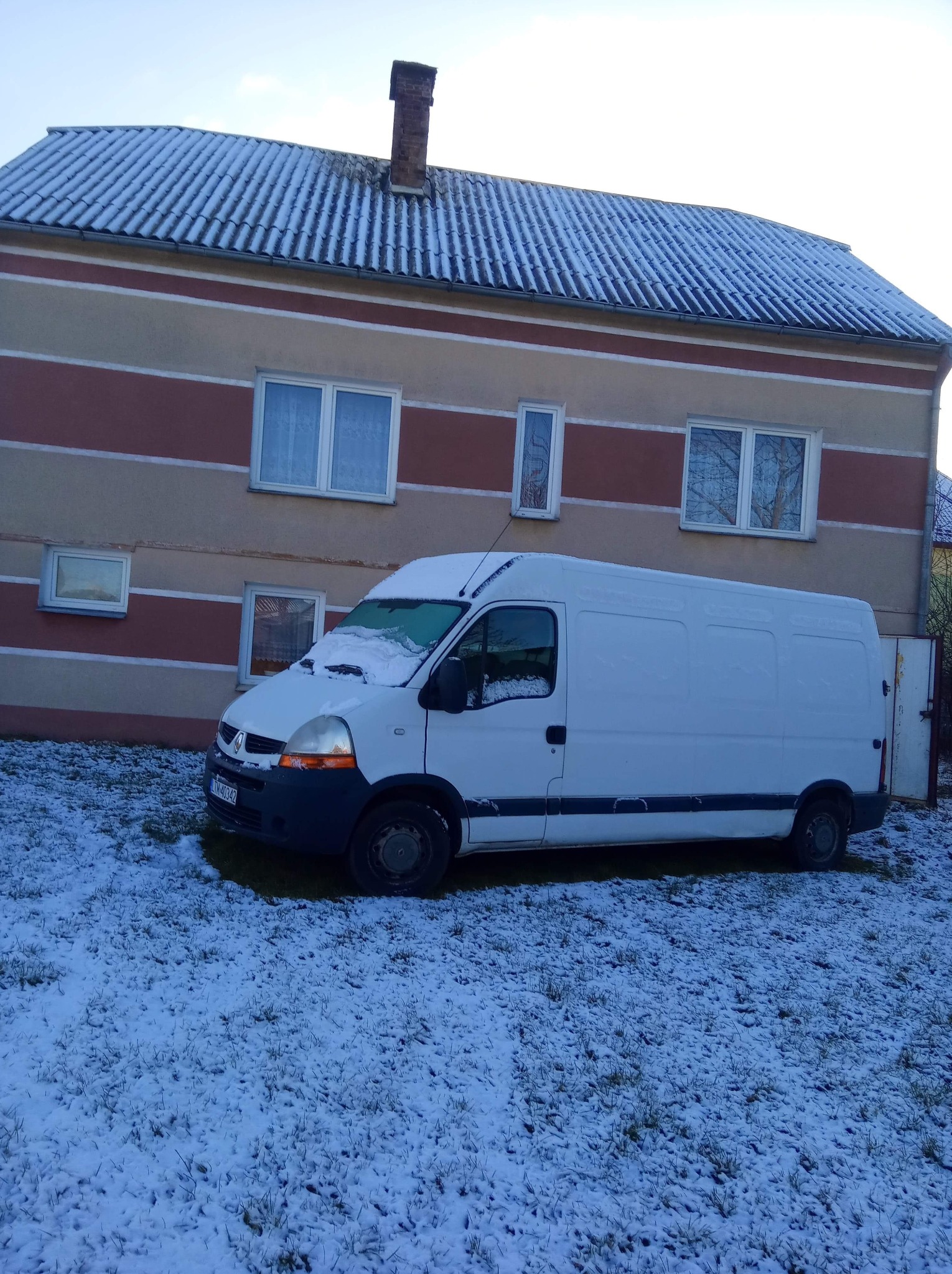 Biały bus dostawczy Renault Master zaparkowany na trawniku pokrytym śniegiem przed dwukondygnacyjnym domem z oknami i kominem.