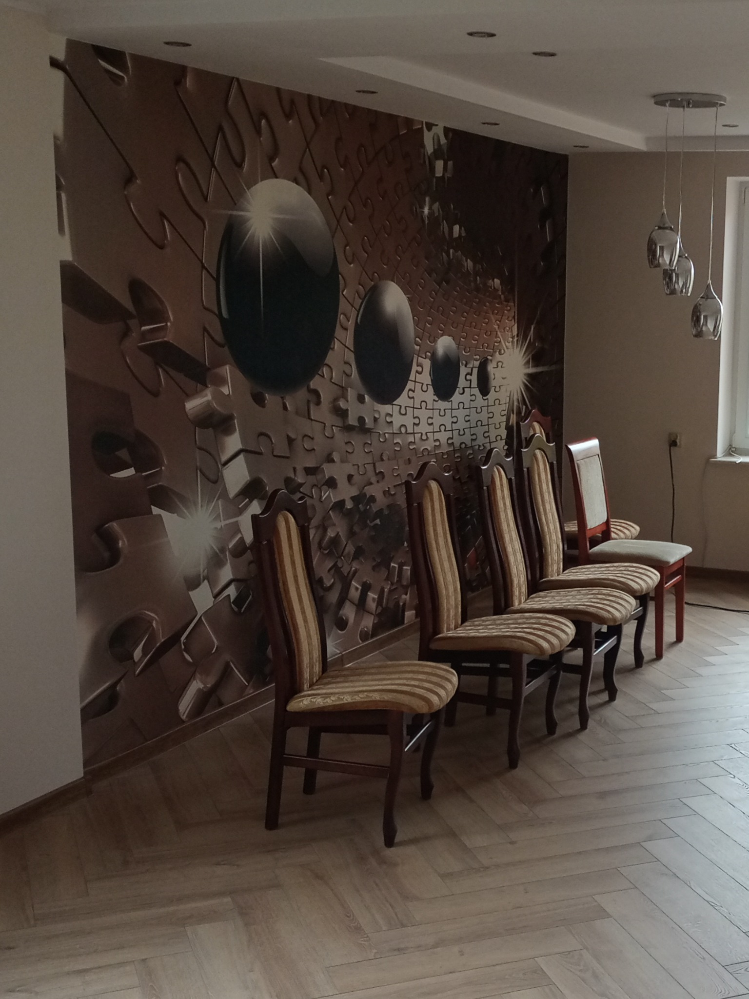 Rząd krzeseł z tapicerowanymi siedziskami ustawiony na parkiecie w jodełkę, na ścianie fototapeta z motywem puzzli i błyszczących kul, nad krzesłami wiszą lampy