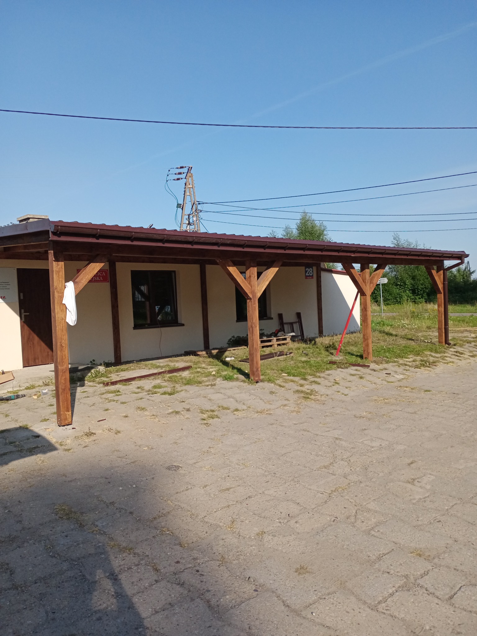 Zadaszenie z brązową blachą i drewnianymi słupami przy budynku z beżową elewacją, widoczne narzędzia i deski, biała szmatka na słupie.