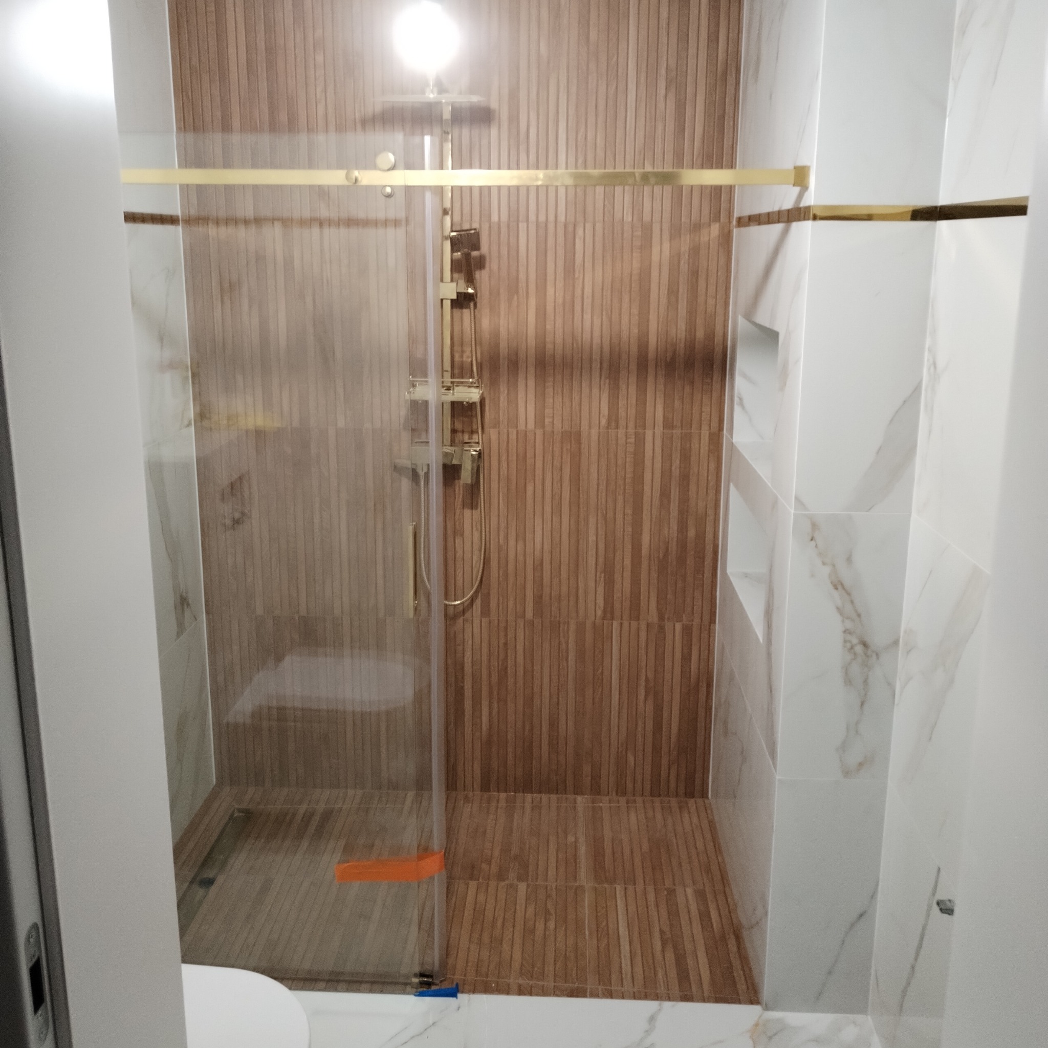 Nowoczesna kabina prysznicowa z elementami w kolorze złotym, drewnopodobne płytki pionowe na ścianie, marmurowe płytki na ścianach bocznych z wnękami.