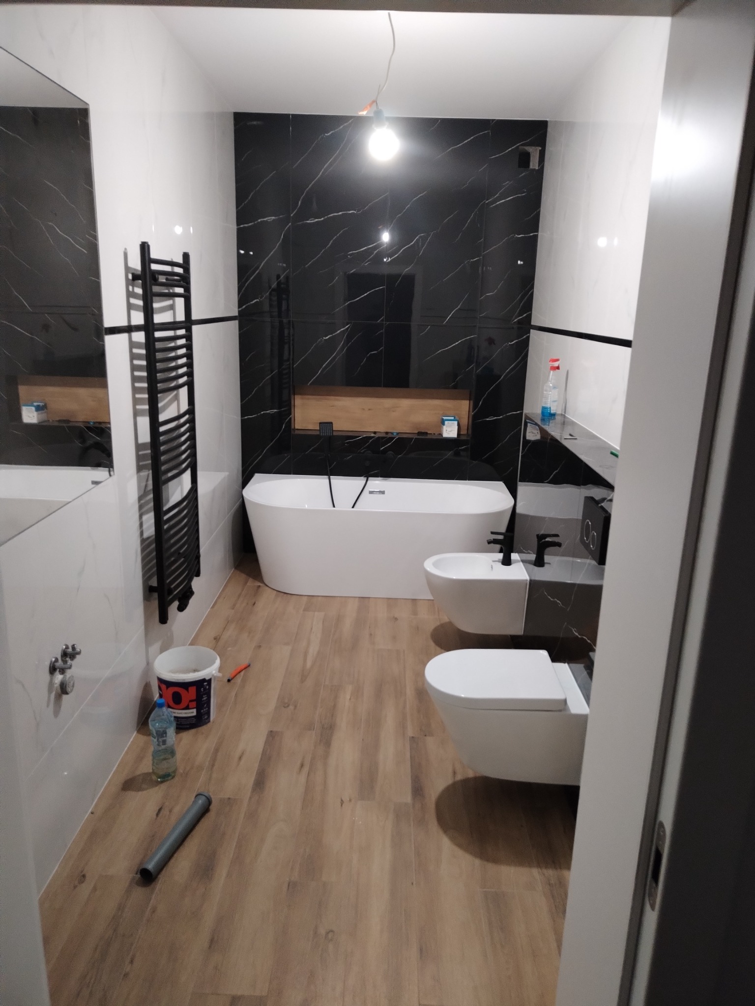 Nowoczesna łazienka z czarno-białymi płytkami imitującymi marmur, wolnostojąca wanna, bidet i toaleta w minimalistycznym stylu, drewnopodobna podłoga, grzejnik drabinkowy i niedokończone detale...