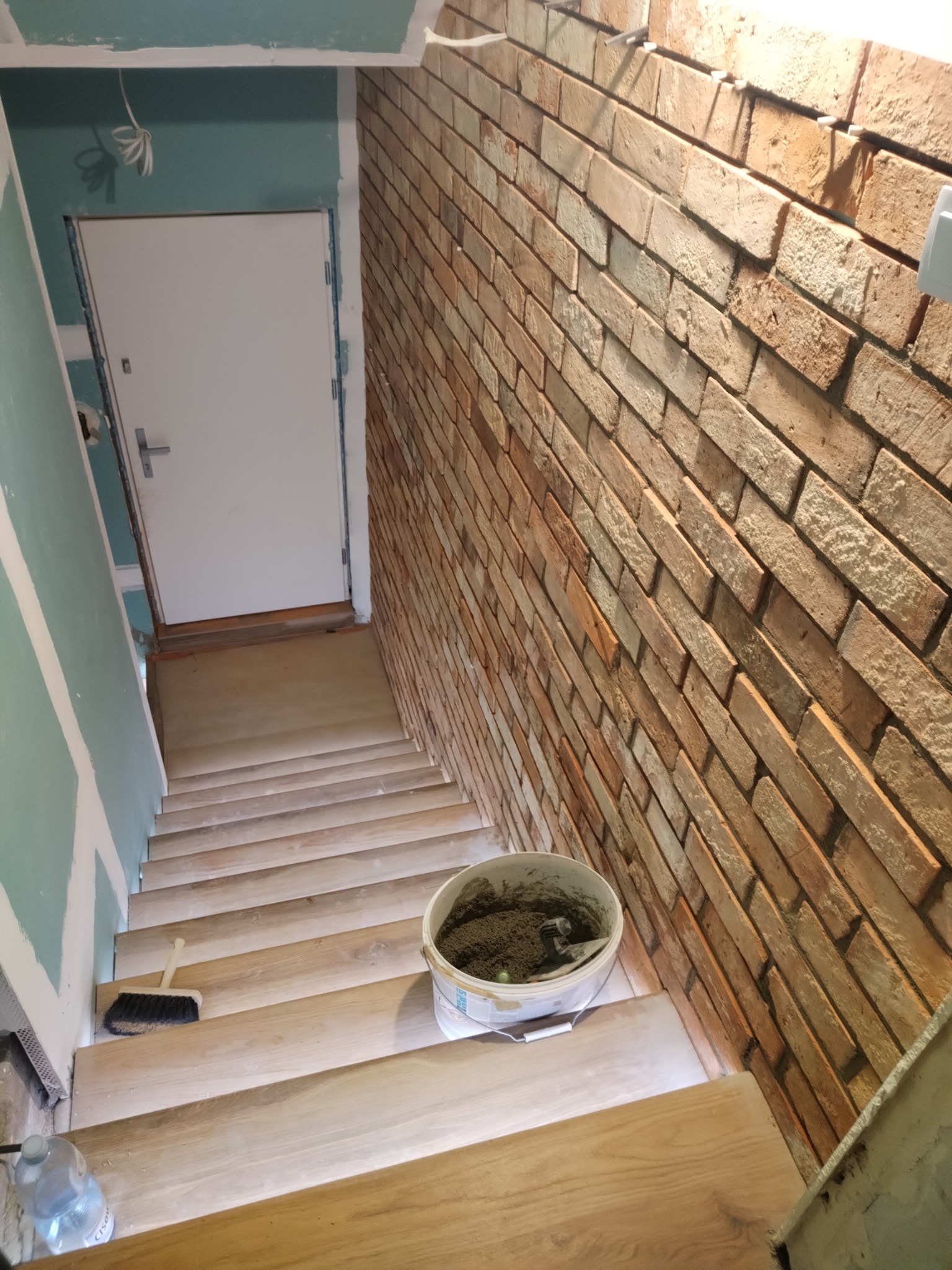 Klatka schodowa w trakcie wykończenia: ściana z cegły klinkierowej, drewniane schody, ściana z płyt karton-gips, wiadro z zaprawą i szczotka.