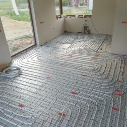 Białe rury ogrzewania podłogowego ułożone na czarnej siatce w jasnym, surowym wnętrzu z dużymi oknami, widoczne narzędzia w tle.