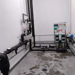 Kompleksowa instalacja rur grzewczych i wodnych z pompami obiegowymi, panelem sterowania i izolacją termiczną, zamontowana w pomieszczeniu technicznym z betonową posadzką.