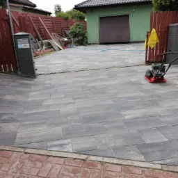 Ułożona szara kostka brukowa na podjeździe, widoczna zagęszczarka w tle, czerwone ogrodzenie z drewnianych desek oraz zielony garaż.