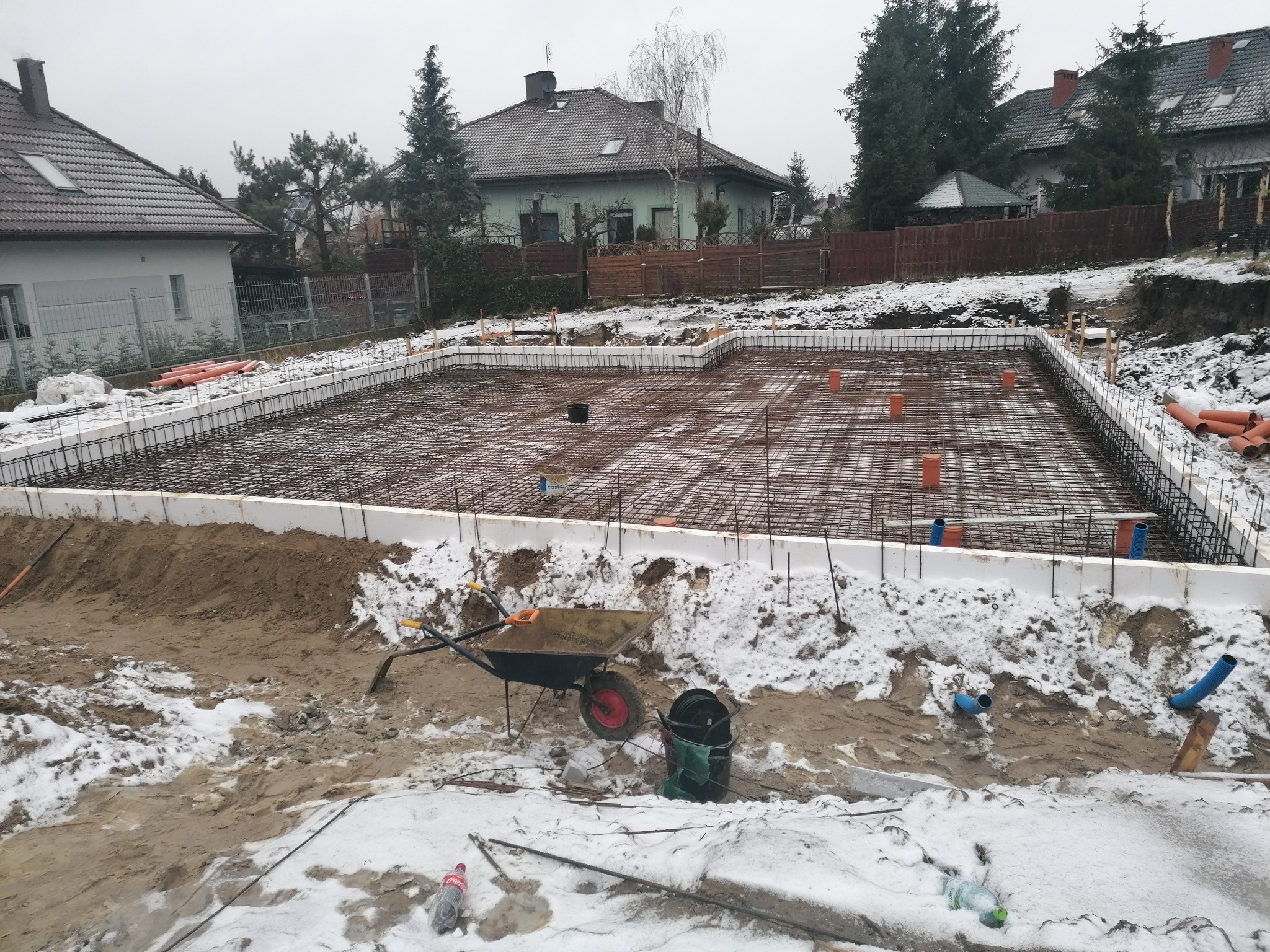 Zbrojenie fundamentów domu jednorodzinnego z użyciem białego szalunku styropianowego, widoczny wózek budowlany, teren pokryty śniegiem, pochmurny dzień.