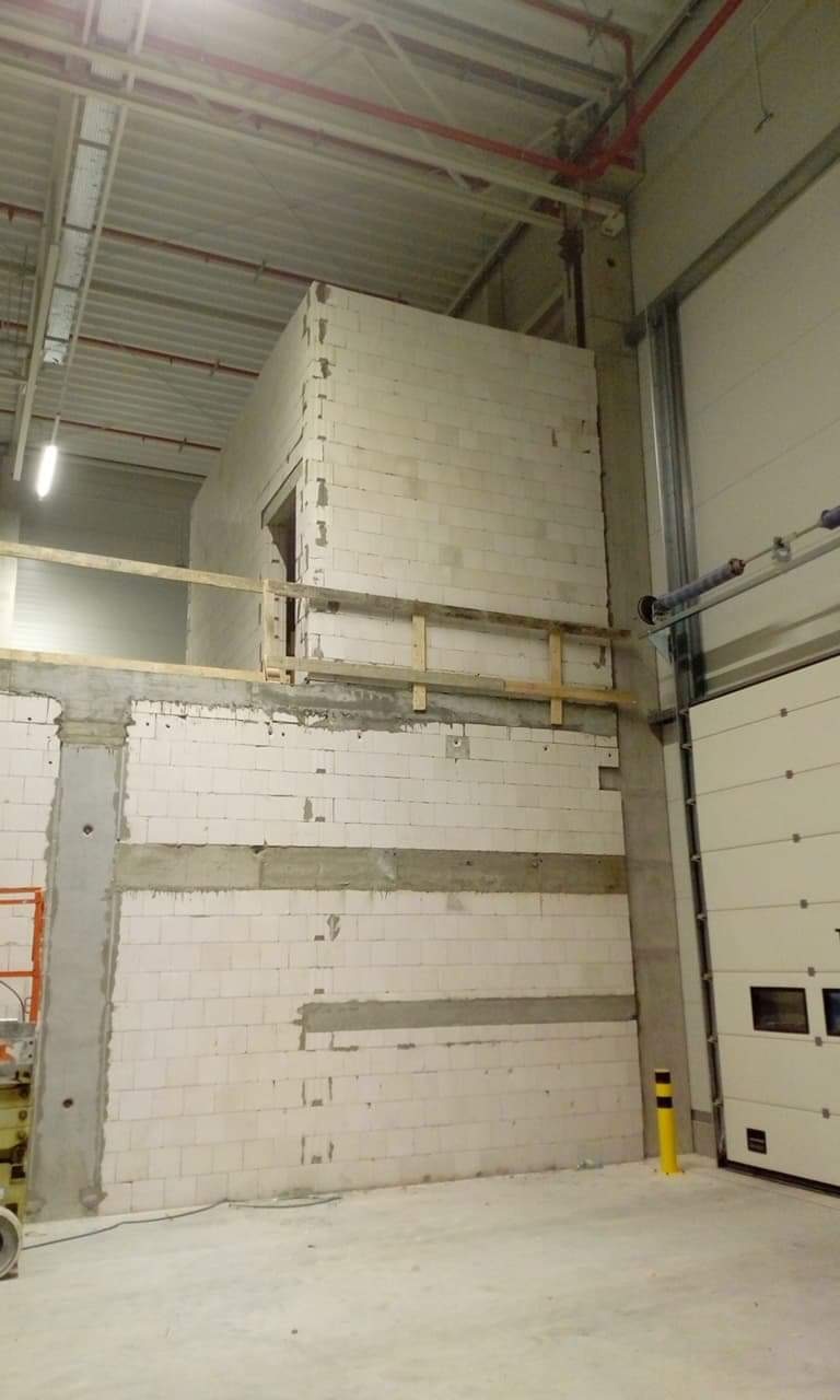 Wznoszenie ścian z białych bloczków wewnątrz hali przemysłowej, widoczne stalowe słupy konstrukcyjne i fragment bramy segmentowej.