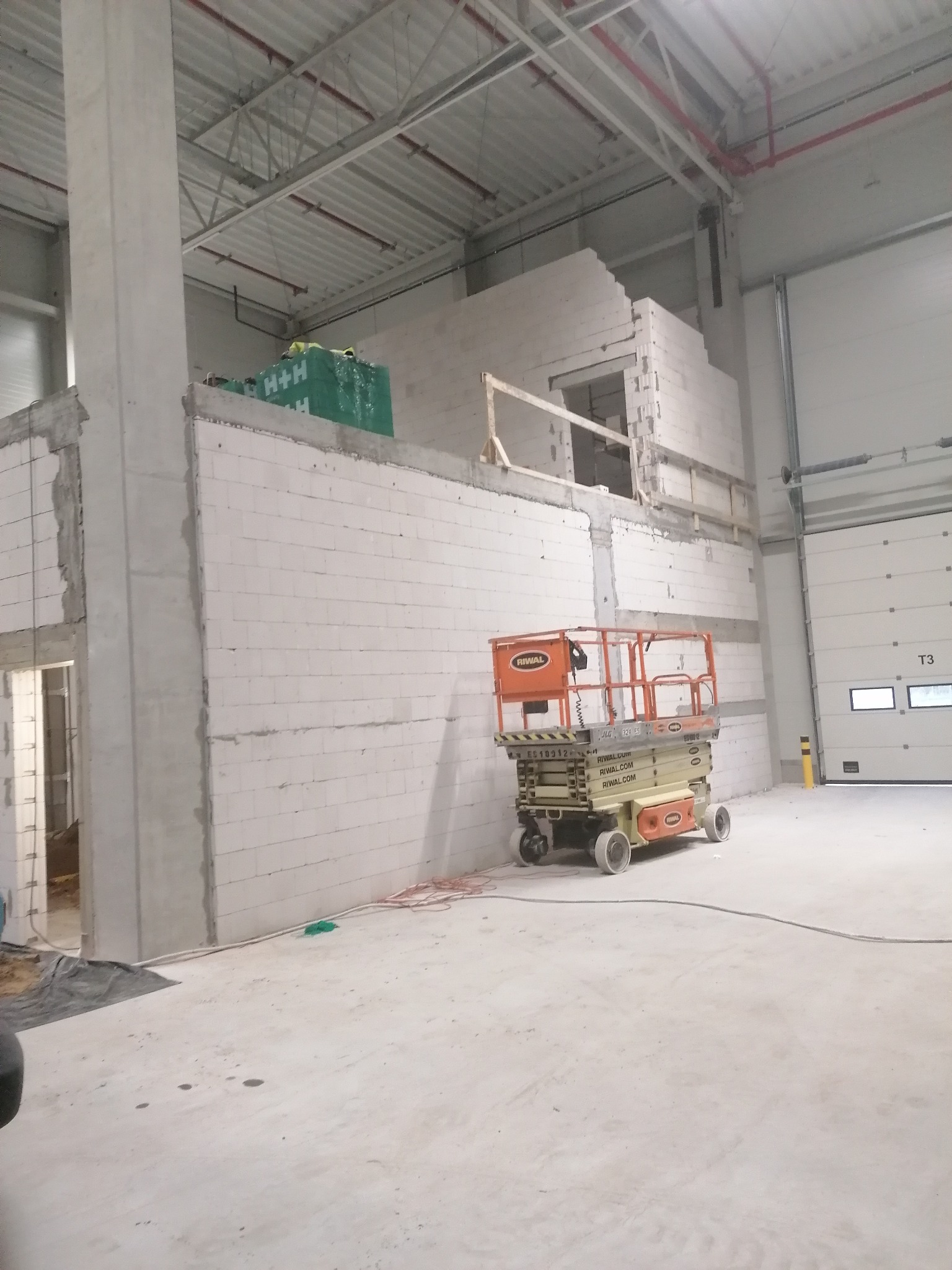 Wnętrze hali w Nowogardzie, w trakcie budowy ścian z bloczków, z podnośnikiem nożycowym Riwal i materiałami budowlanymi na górze ściany, widoczne elementy konstrukcyjne dachu i brama wjazdowa.