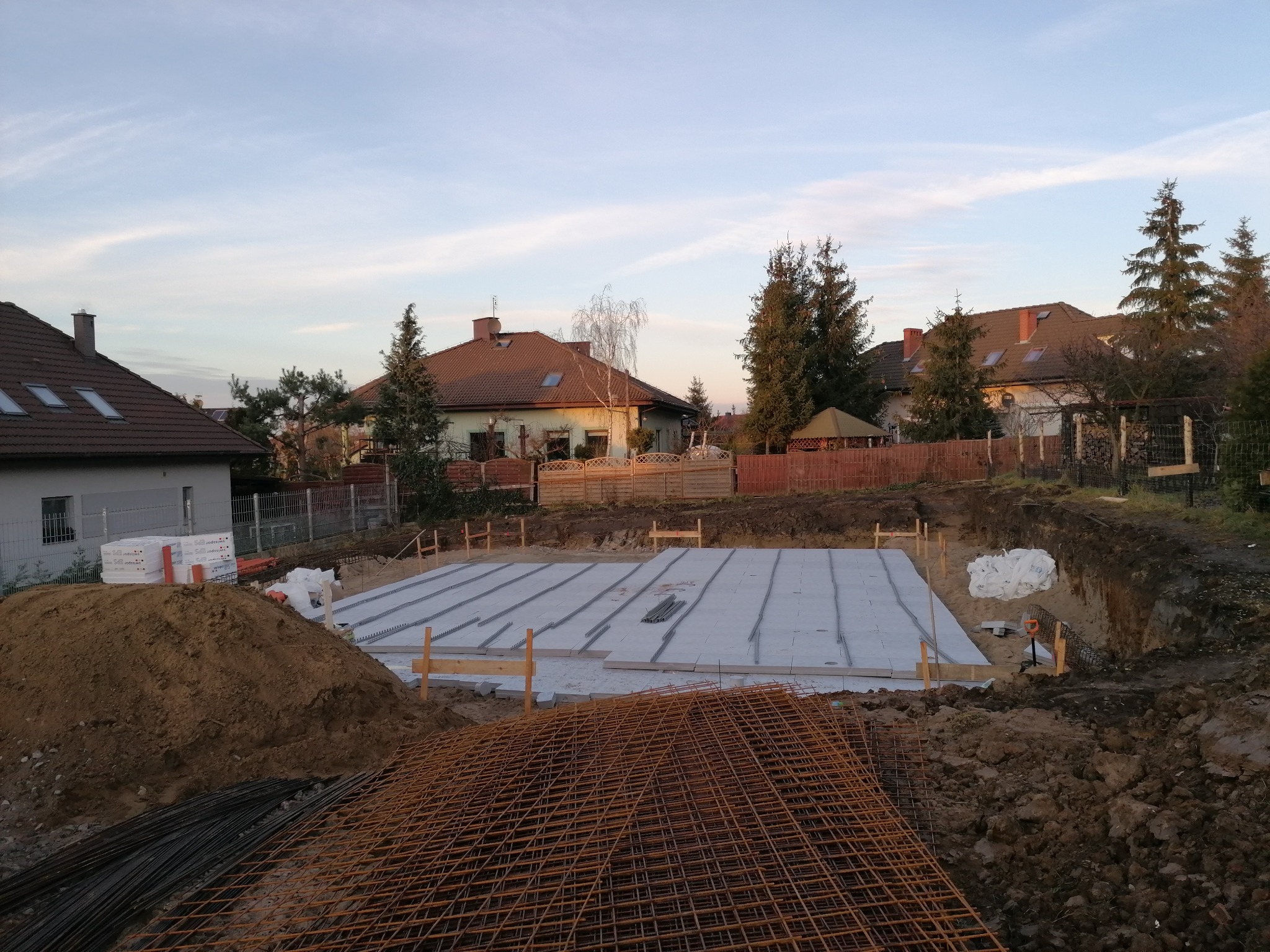 Przygotowany wykop pod fundamenty z ułożonymi bloczkami betonowymi i zbrojeniem w siatce, widok na działkę z domami jednorodzinnymi w tle, wczesny etap budowy.