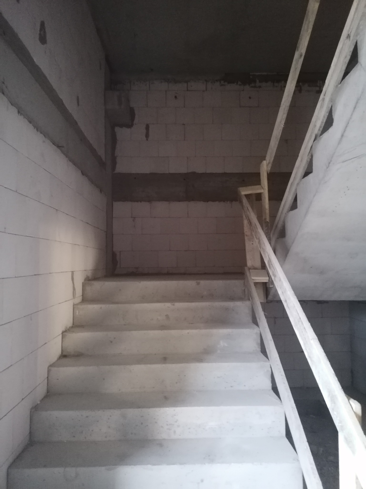 Surowe betonowe schody wewnątrz budynku w trakcie budowy, z prowizoryczną drewnianą barierką, ściany z bloczków betonowych.
