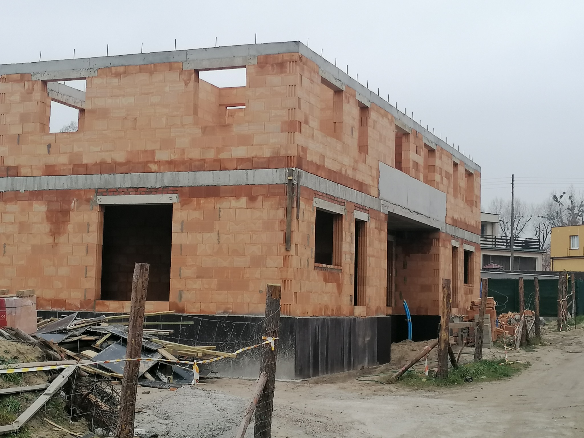 Dwukondygnacyjny budynek w stanie surowym otwartym z czerwonej cegły, z widocznymi otworami okiennymi i elementami betonowymi, teren budowy z pozostałościami materiałów i prowizorycznym ogrodzeniem.