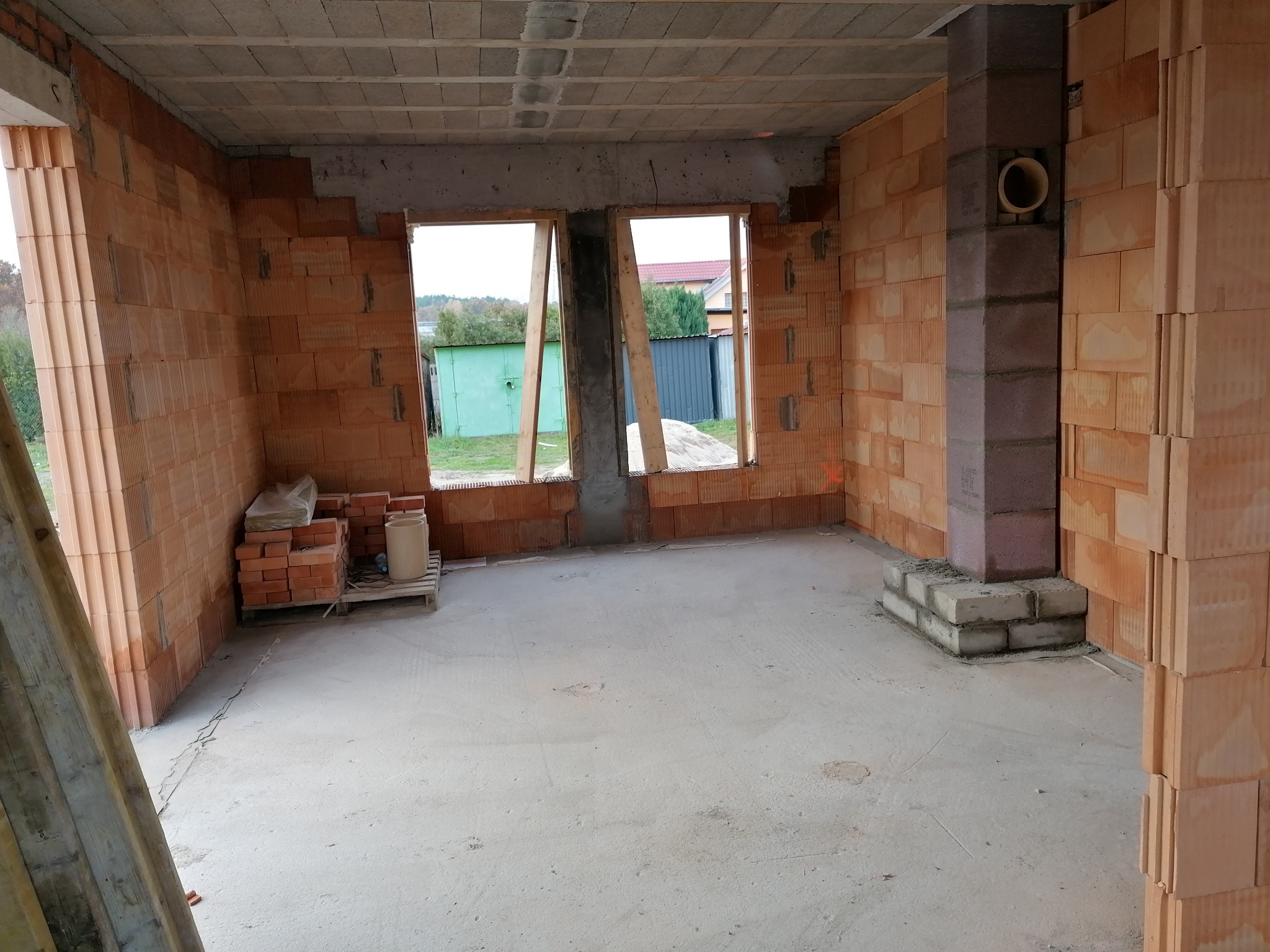 Wnętrze surowego domu w trakcie budowy, ściany z czerwonej cegły, widoczne otwory okienne, betonowa wylewka na podłodze, cegły i paleta w rogu pomieszczenia, komin z otworem wentylacyjnym.