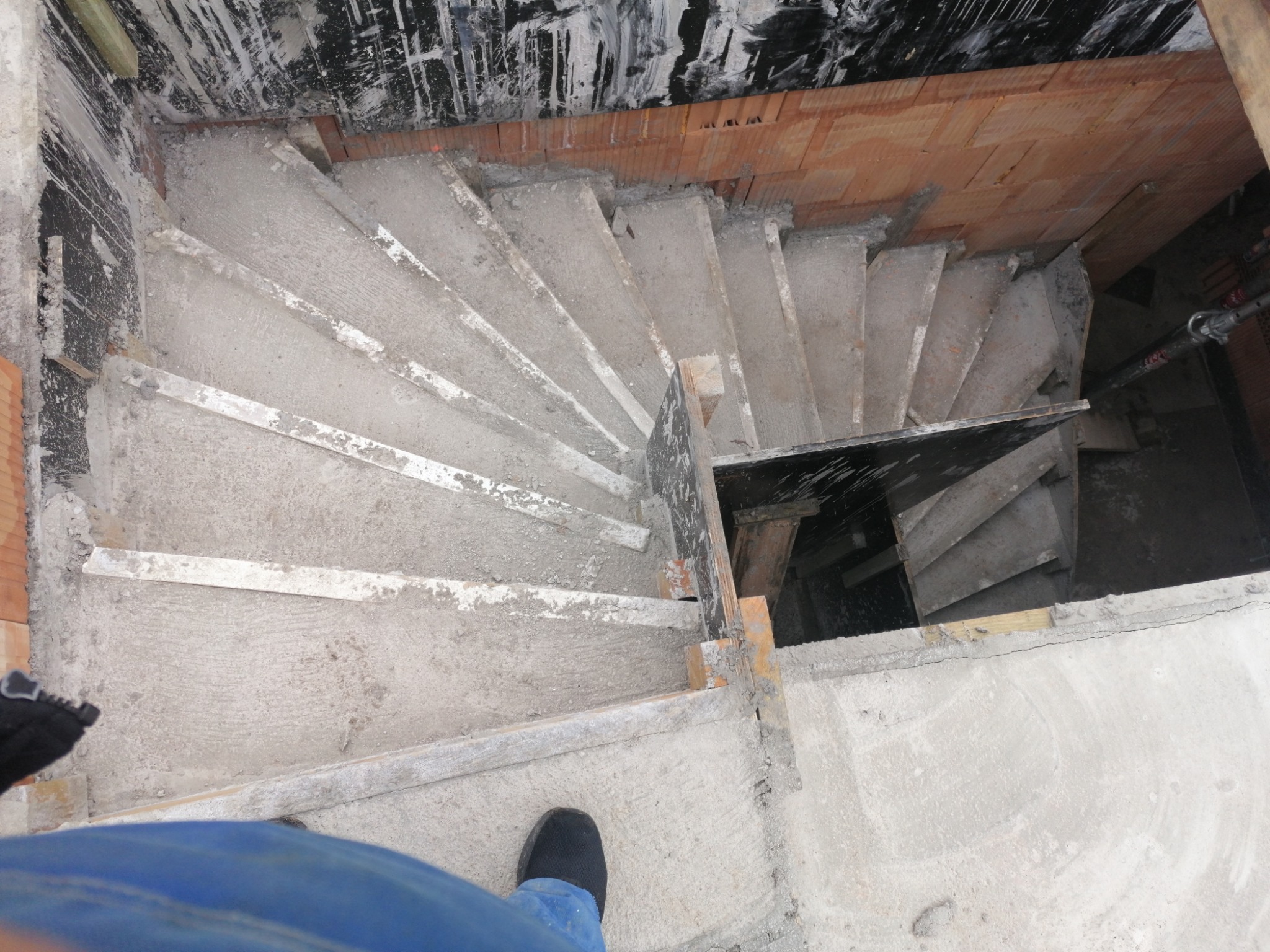 Betonowe schody spiralne w surowym stanie, widok z góry na niedokończoną konstrukcję z szalunkami i fragmentem niebieskich spodni oraz czarnego buta.