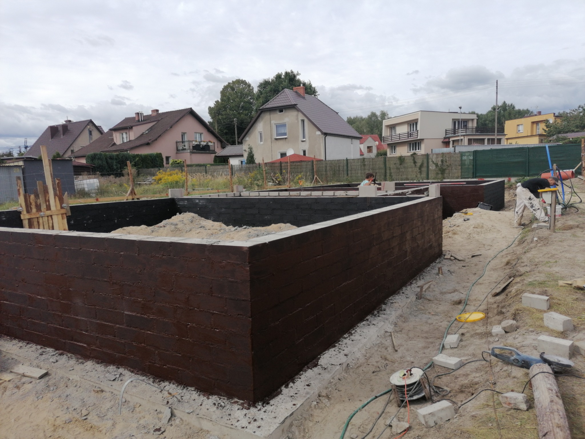 Surowe, brązowe mury fundamentowe w trakcie budowy domu jednorodzinnego, otoczone piaskiem i narzędziami budowlanymi, z widocznymi domami w tle.