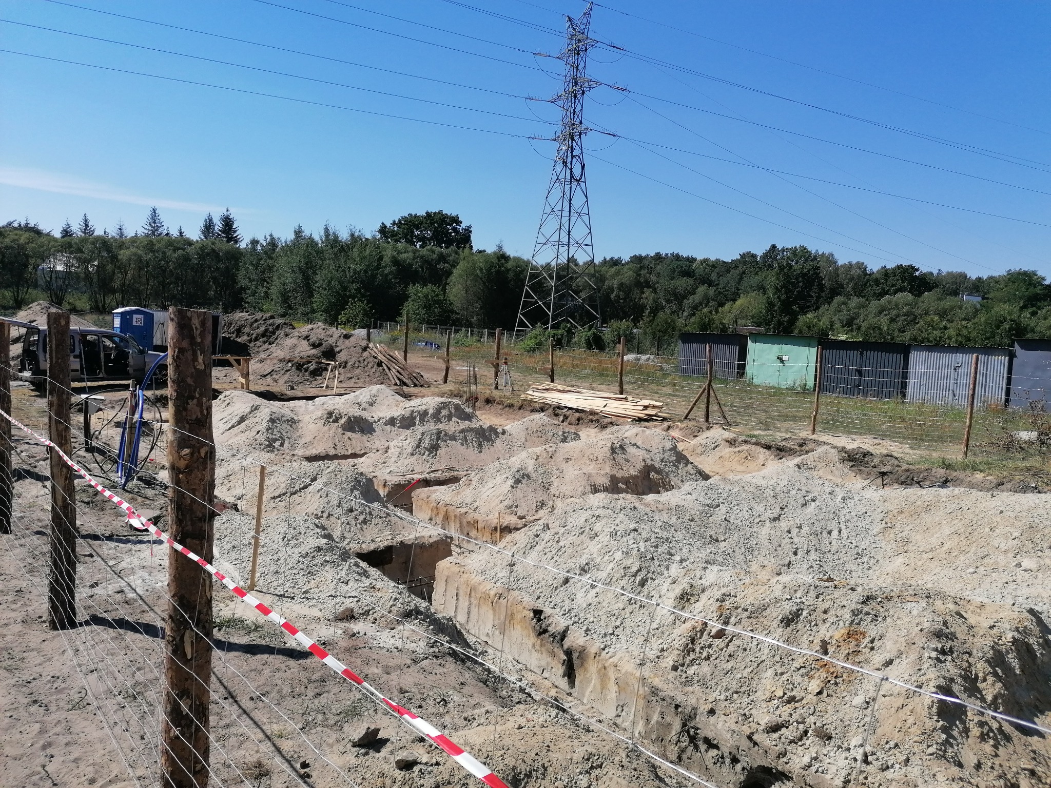 Przygotowanie wykopu pod fundamenty domu jednorodzinnego, widoczne wytyczone rowy i zgromadzona ziemia, w tle słup wysokiego napięcia i zieleń.
