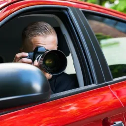 Mężczyzna w czerwonym samochodzie, z krótkimi włosami, robi zdjęcie dużym aparatem z teleobiektywem, wystawiając go przez otwarte okno.