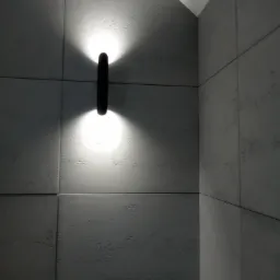 Nowoczesny kinkiet z dwoma punktami świetlnymi, zamontowany na szarej ścianie wyłożonej płytkami imitującymi beton.