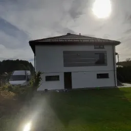 Dwukondygnacyjny dom jednorodzinny z szarą elewacją, brązowym dachem i poziomymi paskami dekoracyjnymi, widoczny z boku, z zaparkowanym białym vanem i puszkami farby na trawniku, w słoneczny dzień.