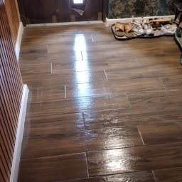 Sokolowski - Świeżo położona, błyszcząca podłoga z płytek imitujących drewno w odcieniach brązu, z widocznym odbiciem światła, listwa przypodłogowa w kolorze białym, fragment drzwi z uszkodzoną okleiną...