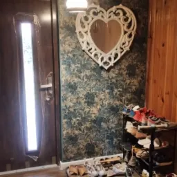 Przedpokój z drewnianą podłogą, wzorzystą tapetą i lustrem w kształcie serca. Obok drzwi stoi regał na buty i hulajnoga.