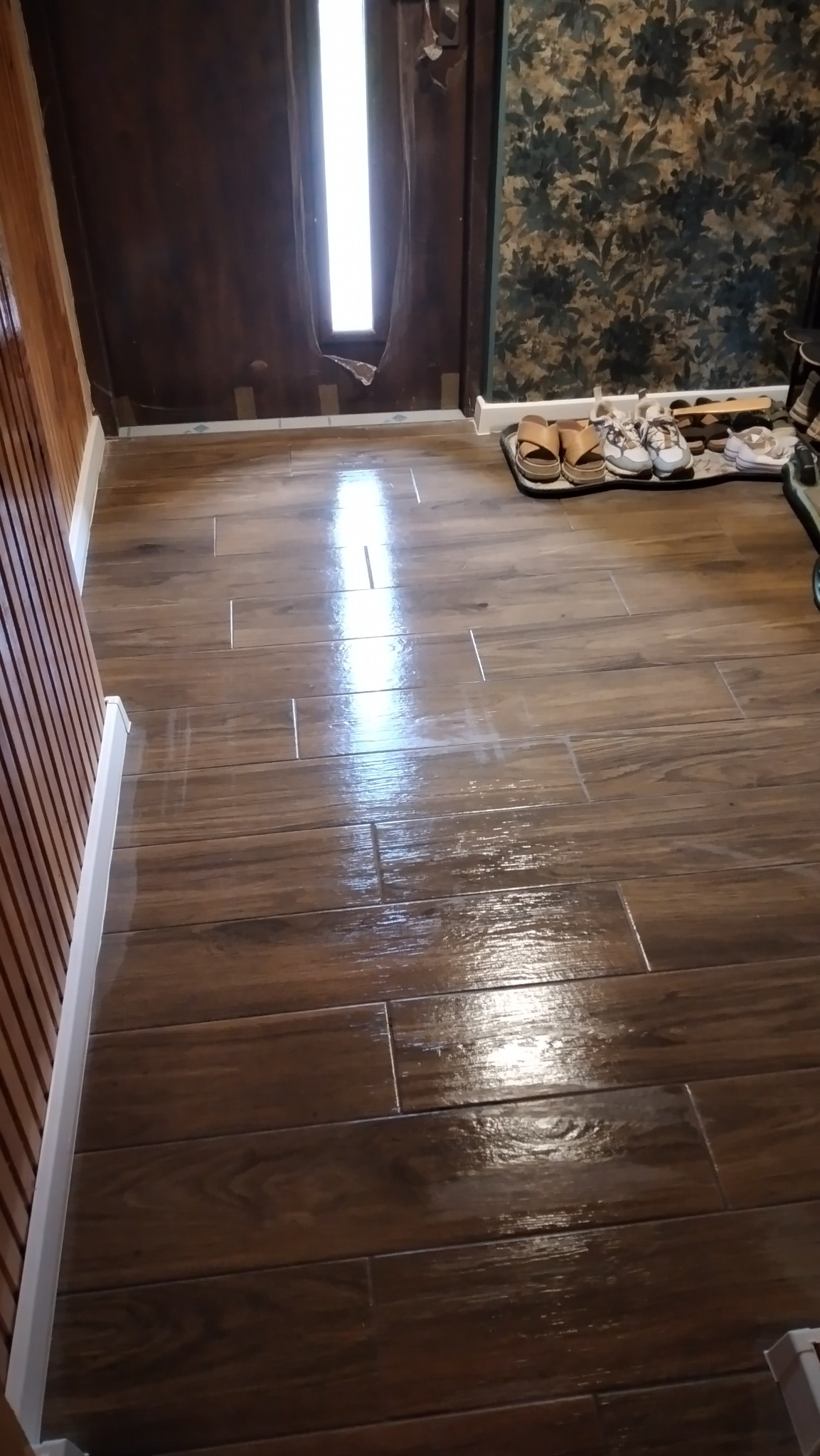 Świeżo położona, błyszcząca podłoga z płytek imitujących drewno w odcieniach brązu, z widocznym odbiciem światła, listwa przypodłogowa w kolorze białym, fragment drzwi z uszkodzoną okleiną...