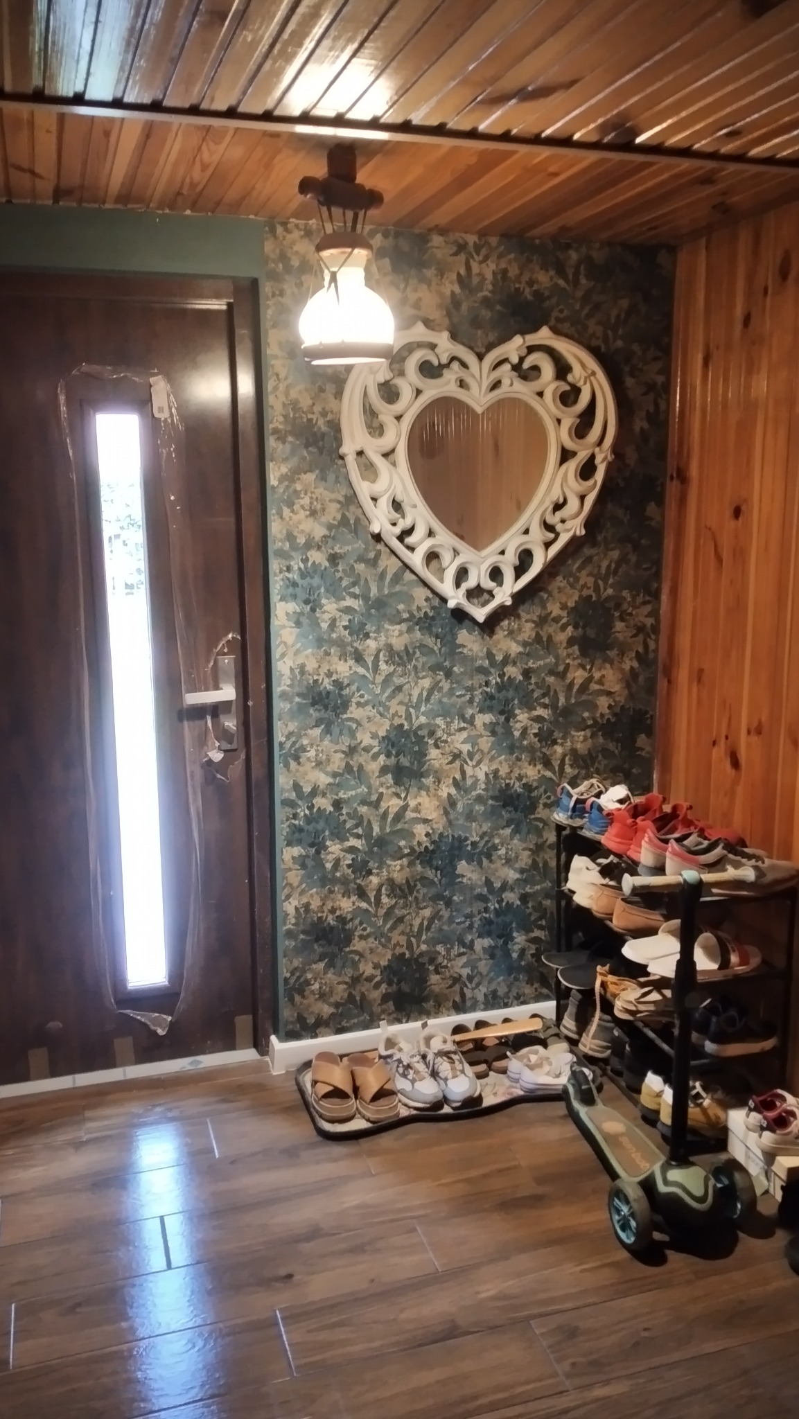 Przedpokój z drewnianą podłogą, wzorzystą tapetą i lustrem w kształcie serca. Obok drzwi stoi regał na buty i hulajnoga.