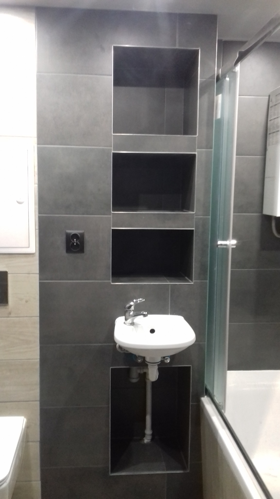 Szara łazienka z umywalką wbudowaną w ścianę z wnękami, widoczna kabina prysznicowa i biała ceramika.