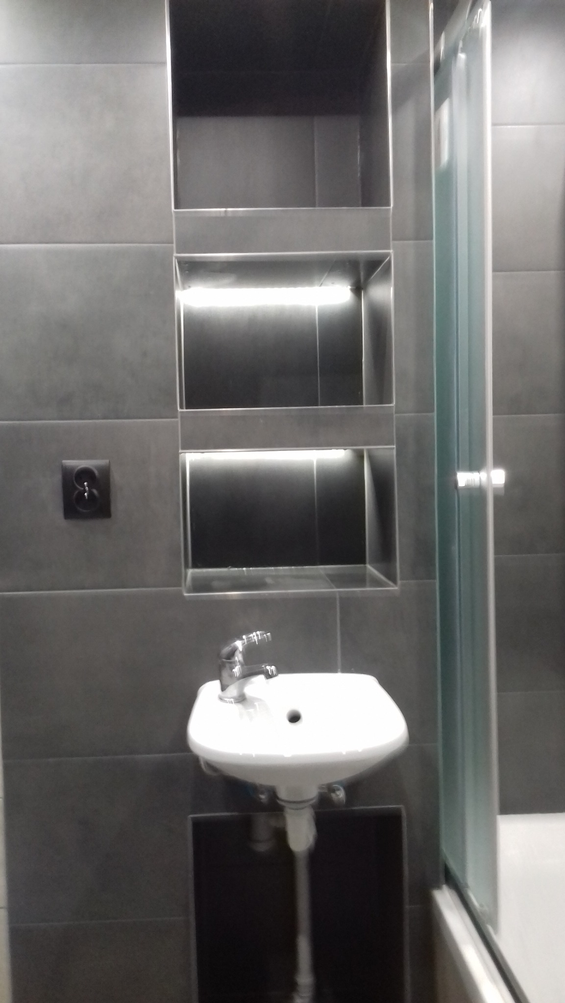 Nowoczesna łazienka z szarą glazurą, podświetlanymi wnękami i białą umywalką nablatową na czarnej szafce, widoczny fragment kabiny prysznicowej.